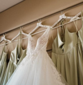 A white lace wedding dress hanging on a white hanger next to sage green satin bridesmaid dresses on a wall-mounted display.