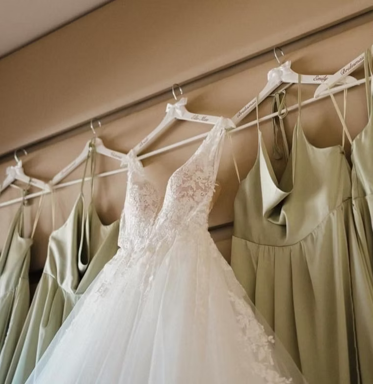 A white lace wedding dress hanging on a white hanger next to sage green satin bridesmaid dresses on a wall-mounted display.